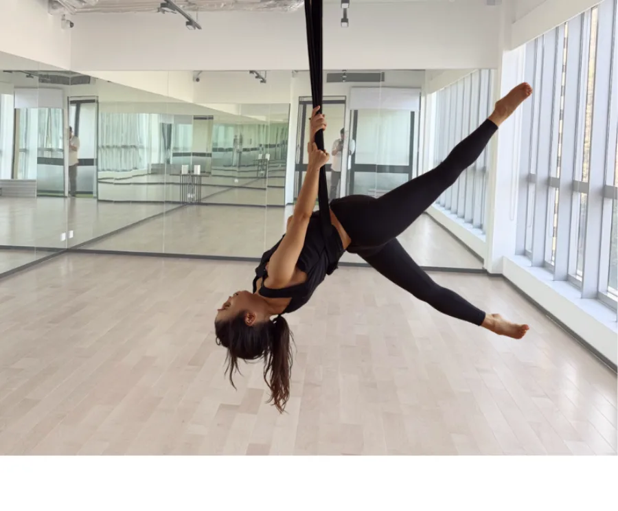 An adult hanging upside down from a bungee strap with legs in a wide split, in the mirrored studio at MosKiddo