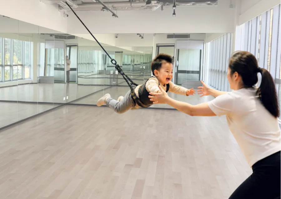 A toddler in a bungee harness reaching for an instructor's hands in the studio at MosKiddo