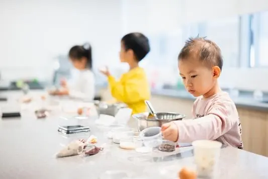 A young child mixing ingredients in a bowl at the MosKiddo baking bench, with two others behind